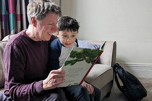 James on a sofa in a comfortable room, reading a book with a young child. Both James and the child look really excited at what they're reading.