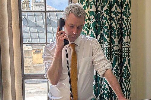 Will Forster speaking on the phone in his Westminster office, he is wearing a white shirt with the sleeves rolled up and a yellow tie.