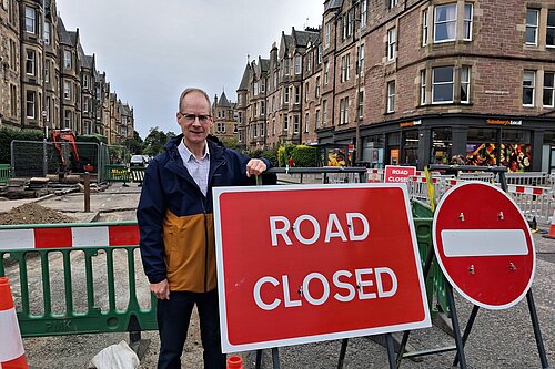 Councillor Neil Ross standing next to a "Road Closed" sign with buildings in the background
