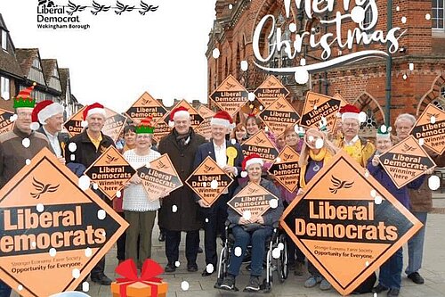 Lib dems by Wokingham town hall with Santa hats etc superimposed on top. The Wokingham Lib Dem logo has been made to look like Santa’s sleigh being pulled by Lib Dem