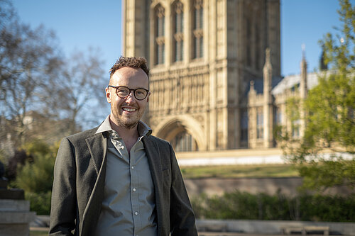 A man wearing glasses stands smiling outdoors in front of a large historic building.