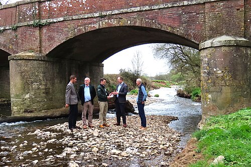 A photo of a group of people standing on a rocky exposed section of riverbed by an old stone bridge.
