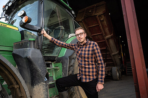 Adam Dance MP standing next to a large green tractor inside a barn, wearing a checked shirt and black trousers, with one hand holding the vehicle door and one foot resting on the step.