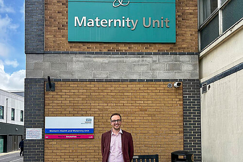 A man stands outside a hospital building under a sign for the maternity unit.