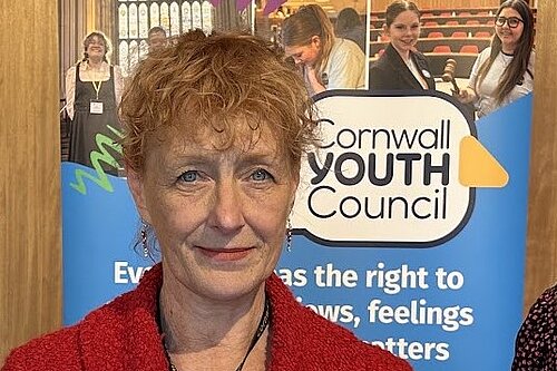 A woman with short red hair stands in front of a poster. Text of the poster reads Cornwall Youth Council.