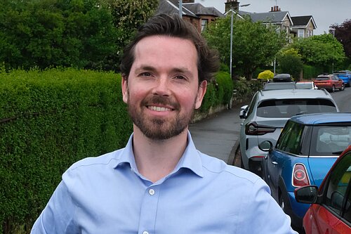 Adam Harley standing smiling with cars parked on the road next to the pavement behind