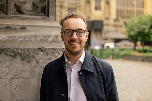 A man stands in a shirt and a black coat, wearing glasses with a historic building in the background.