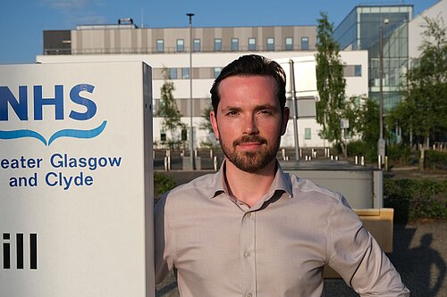 Adam Harley next to a sign with the NHS Greater Glasgow and Clyde logo with a hospital building in the background