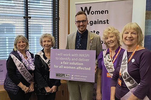 Adam Dance MP stands with four WASPI campaigners, all smiling and holding a purple sign that reads, “I will work with WASPI to identify and deliver a fair solution for all women affected.”