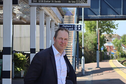 A photo of Richard Foord MP standing on the platform in front of the ticket office at Honiton Station on a sunny day