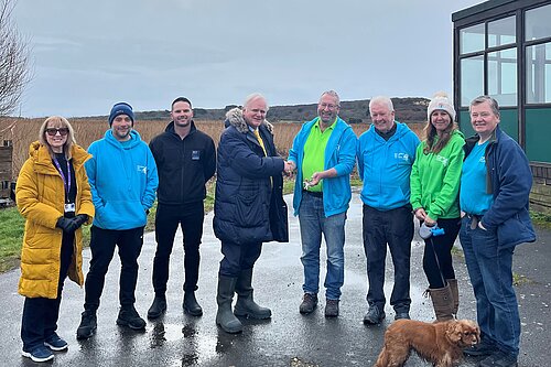 Members of Hengistbury Head Outdoors with Councillor Mike Cox and the BCP Council team posing in a line outside the centre. There are two women and six men in the group, and a brown spaniel. It's a grey, wet day and five of the people are wearing matching brightly-coloured blue or green hoodies