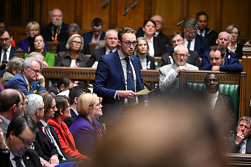 A man in a suit stands speaking in the House of Commons chamber, holding a piece of paper. Other MPs sit and listen around him.