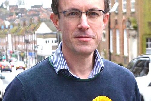 Close up of Ben with LibDem rosette, looking at the camera, with Lewes's high street in tha background