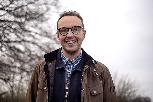 Portrait of a man wearing glasses and a brown coat, with a tree in the background. 