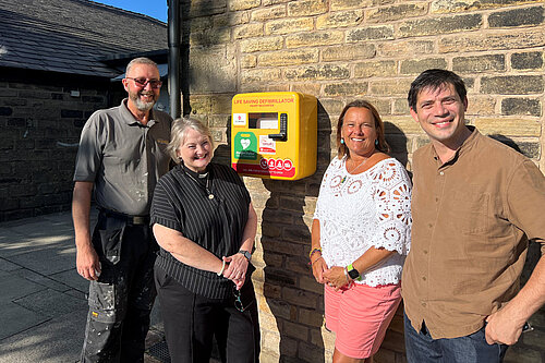 Church Warden Steven Reed, Vicar of Leesfield and Hey Reverend Kirsten Stott, Councillor Alicia Marland, Councillor Sam Al Hamdani at St John's Church with the defibrillator